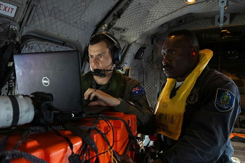 Two military men, one wearing a life vest, look at a screen while inside an aircraft.