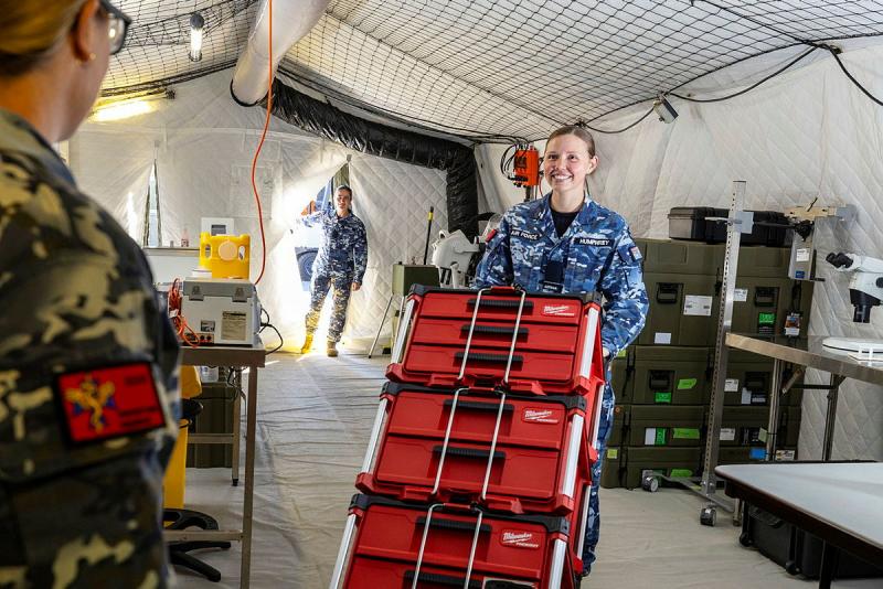 A woman in an Air Force uniform wheels a stack of red equipment boxes as two others look on.