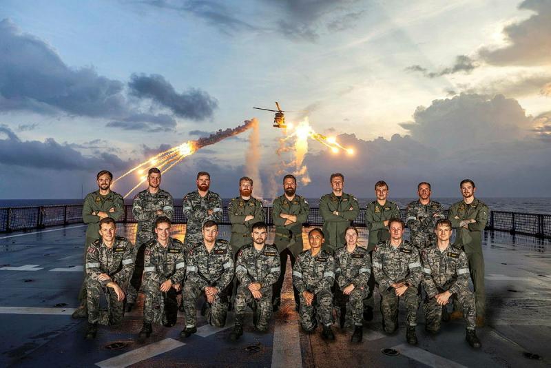 A group poses on a ship while a helicopter fires flares in the background.