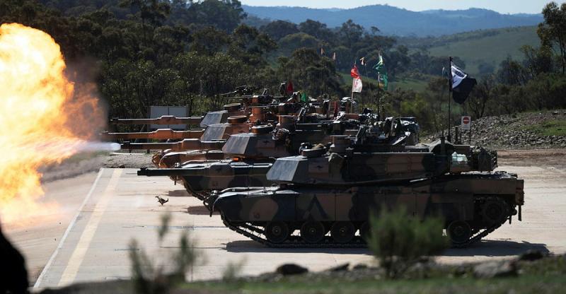 A small group of tanks line up and fire on a concrete clearing in a bush setting.