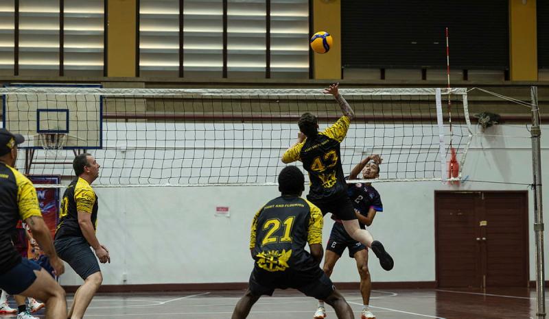 Two teams face each other on a volleyball court with one person jumping up high to hit the ball.