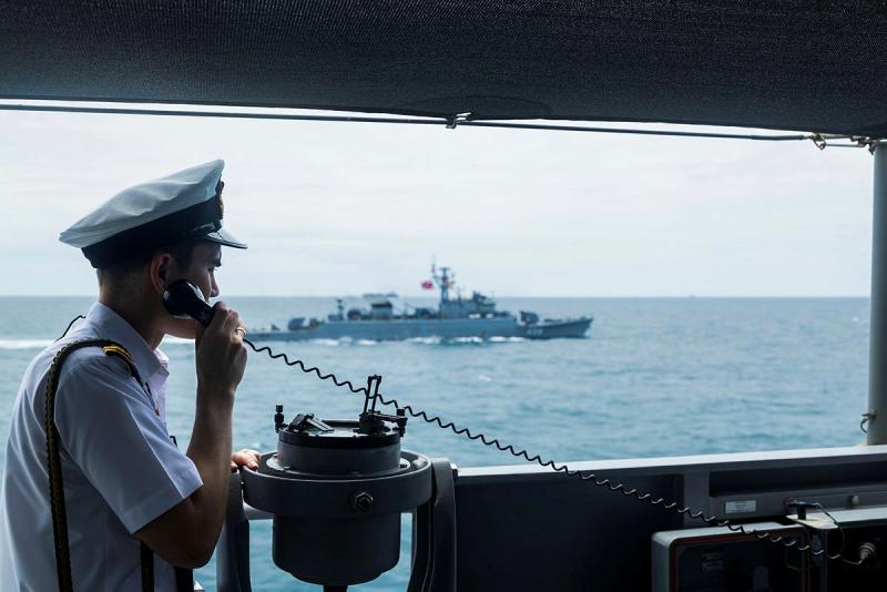 A Navy member stands holding a handset to his ear on board a ship while another is seen in the background.