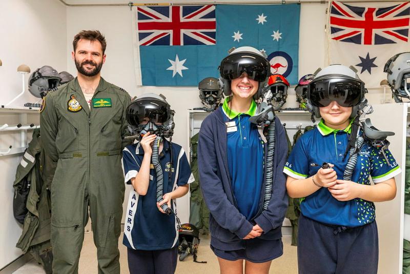 Three students try on pilot helmets while next to a military man.