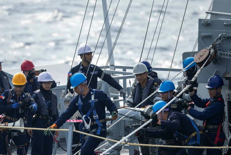 International Navy personnel stand on the deck of s ship wearing hard hats in varying colours.