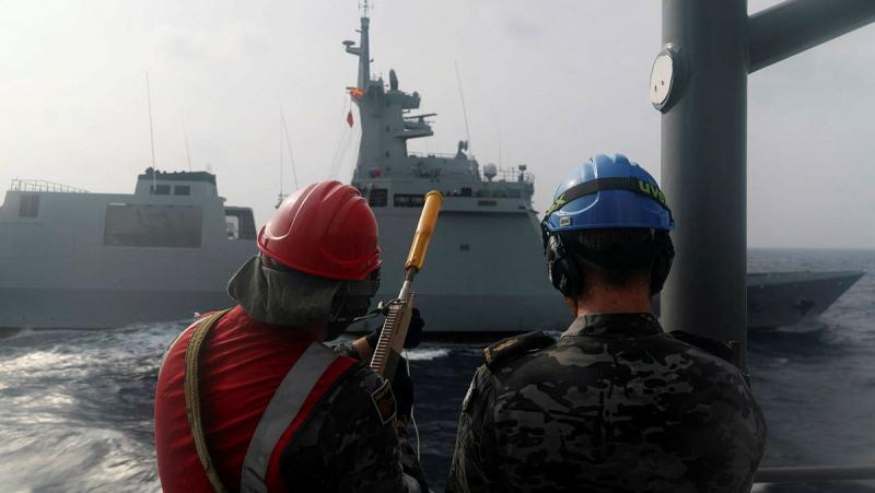 Two personnel in hard hats stand ready to fire a line to another ship from theirs.