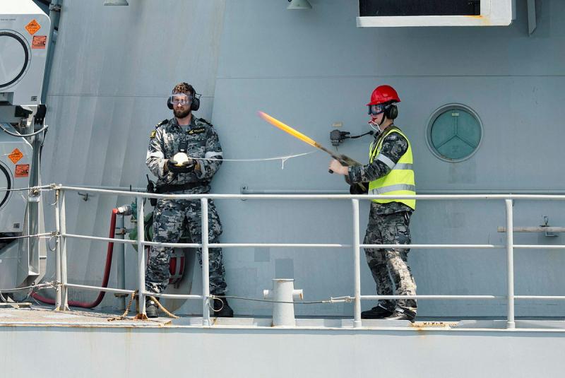 Two Navy personnel stand together, one aiming a gun out to sea and the other holding a line.