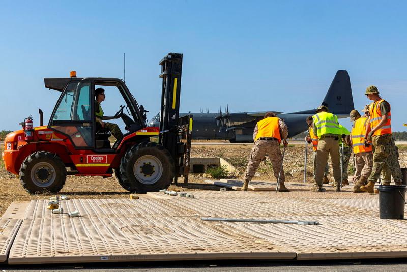 Several military members in high-vis lay mats onto the ground, with an aircraft in the background and the help of a small digger.