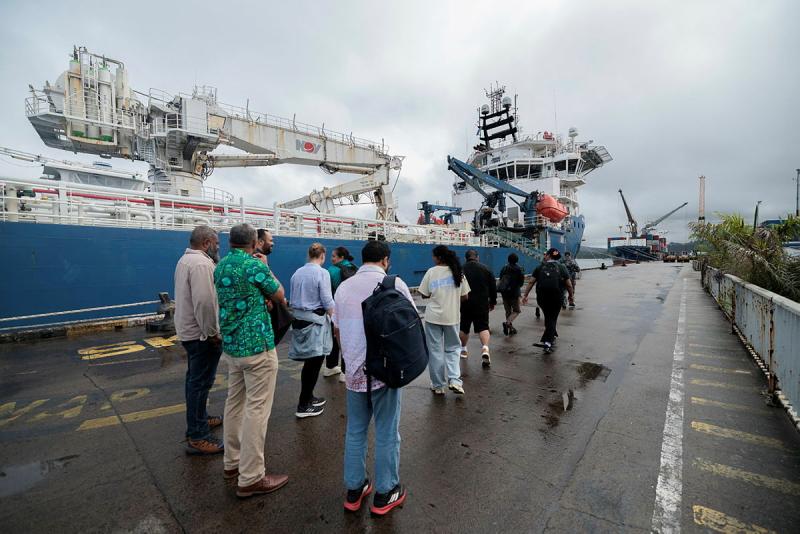 A line of people are seen standing in a concrete area next to a ship on a cloudy day.