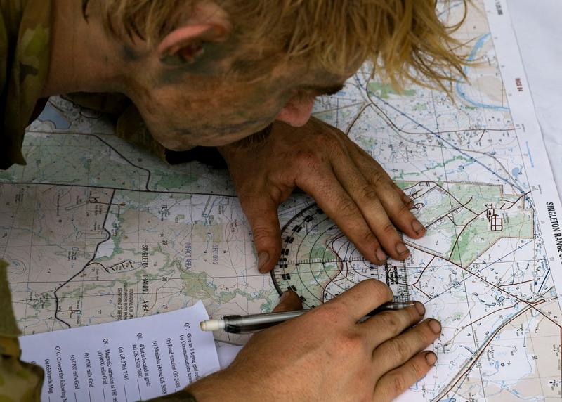 A man with camo paint on his face uses a measuring device on a map.