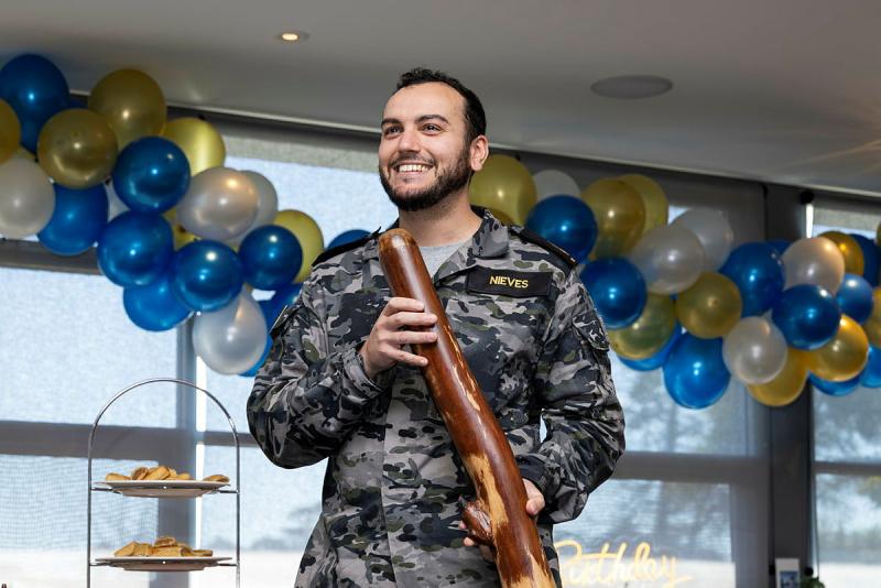 A sailor holds a didgeridoo in front of balloons.