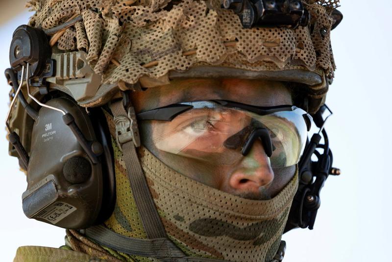 A close-up shot of a military man wearing glasses, a headphone set and a covering over his head and mouth.
