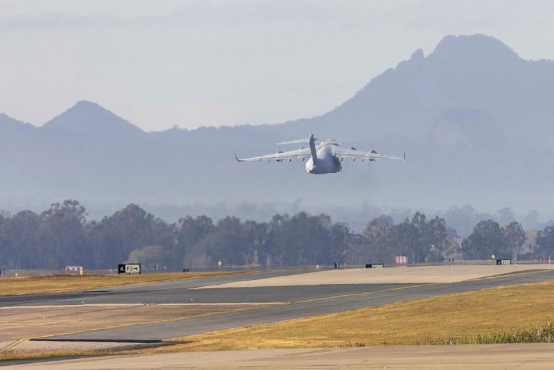 An aircraft flies low over the tarmac and towards a mountain region.