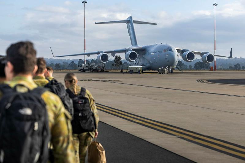 Military personnel with backpacks walk across the tarmac to a stationary aircraft.