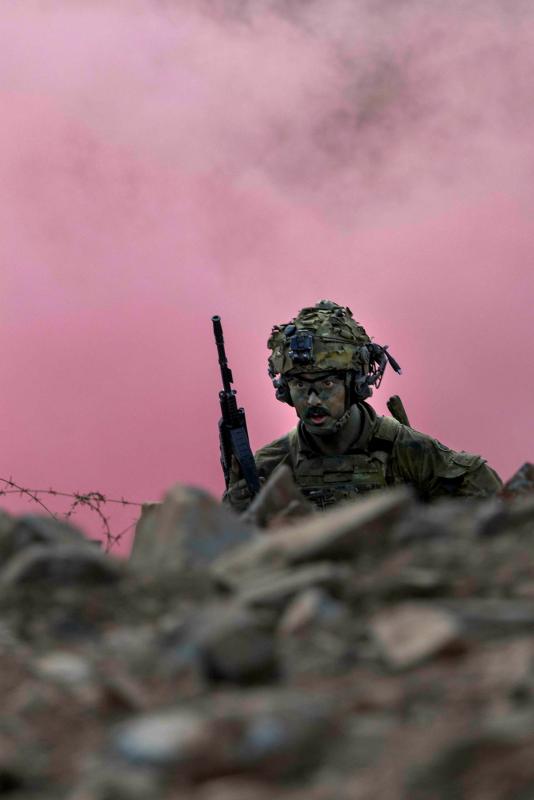 An armed soldier runs around barbed wire and rocks with pink smoke in the air.
