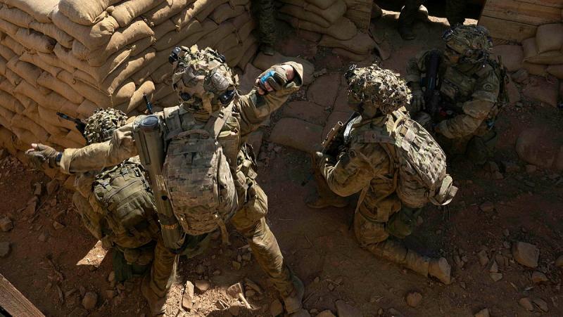 Several military members crouch behind a trench wall; one of them throws something like a ball.