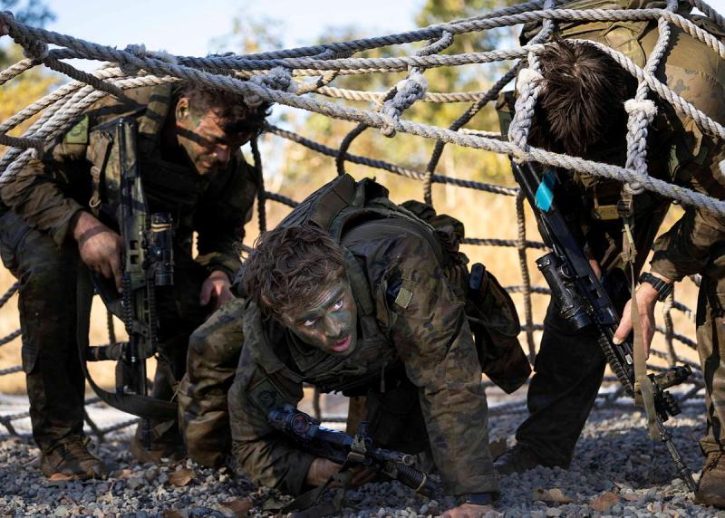 Three armed military members crawl beneath a large rope net.