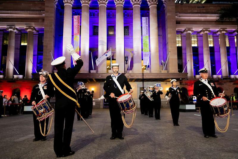 Navy members play instruments and drums in front of a building with concrete columns lit from behind with a purple light.