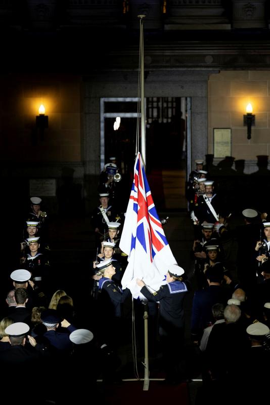A white, blue and red flag falls around a flagpole as people in black and white uniforms look on in a darkened rom.
