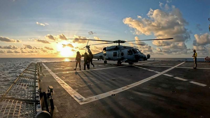 With the sun low in the sky, golden light shines on a helicopter and three people on a ship at sea.