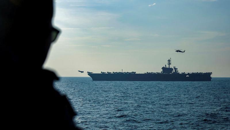 A person seen in silhouette looks out to sea, where aircraft take off from a large vessel.
