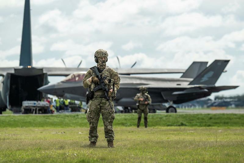 Two armed military members guard several aircraft on the tarmac.