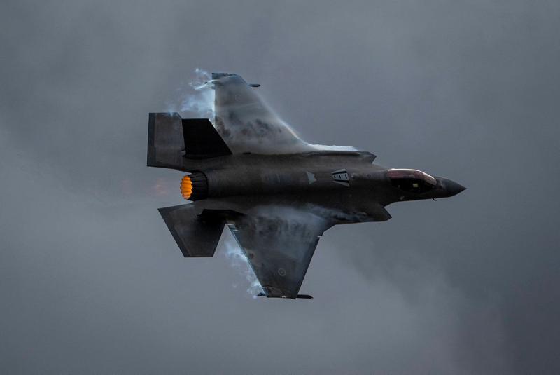 Mist streams off the wings of an aircraft at flight in a dark grey sky.