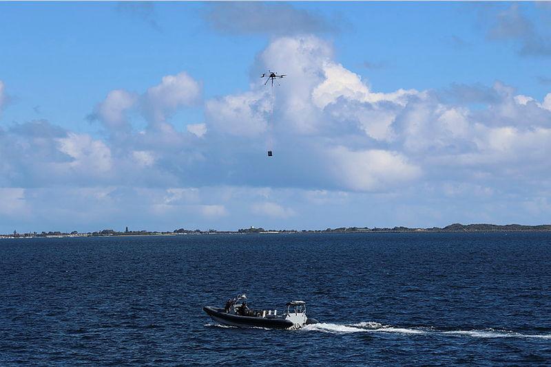 A drone carrying a load on a cable flies over water, above a boat.