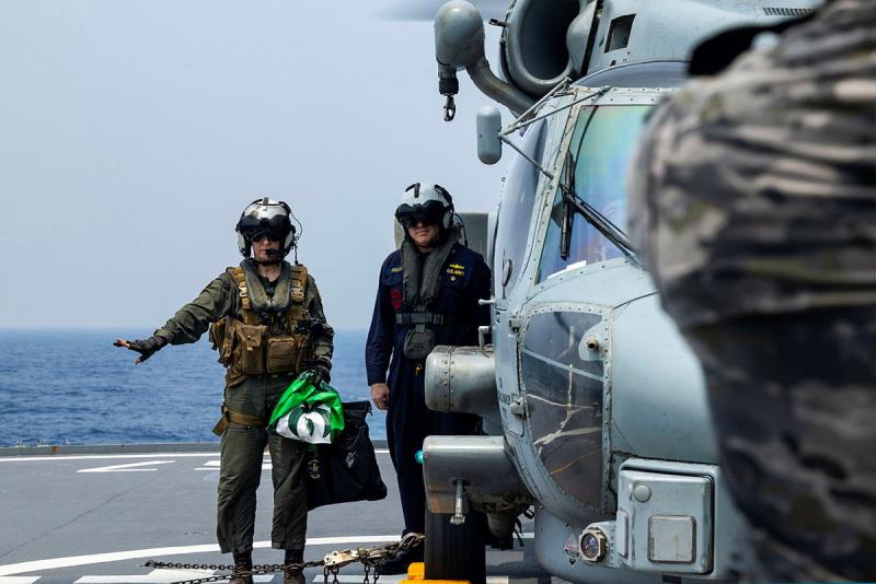 Two Navy personnel stand next to a helicopter on the flight deck of a ship.