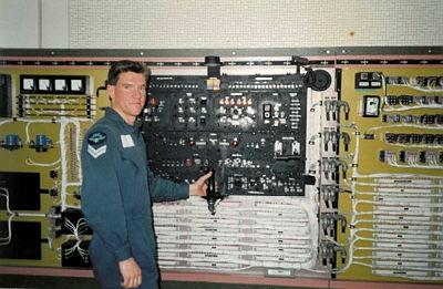 A man in blue coveralls checks electrical equipment and looks at the camera.