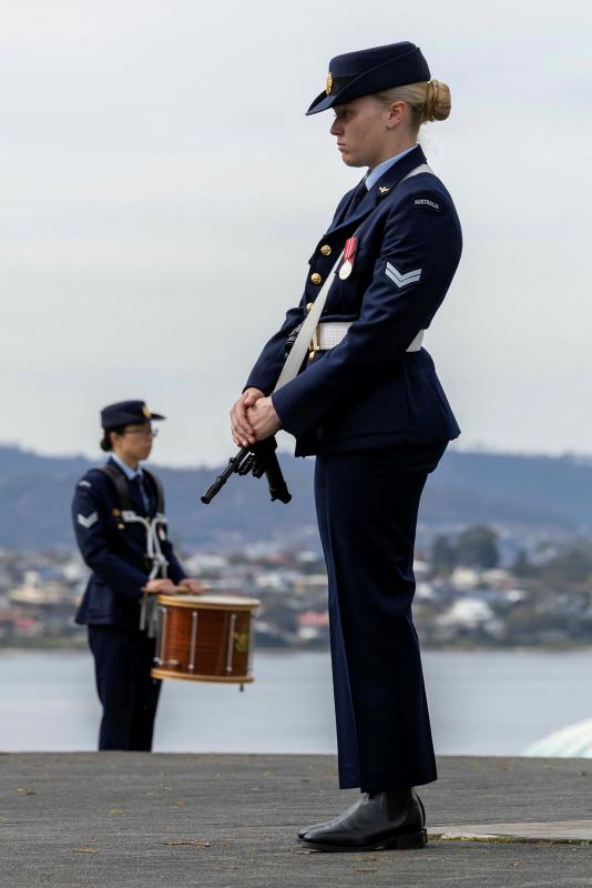 A military woman stands with her hands on her gun and her head bowed, as another military woman holds a drum to her left.