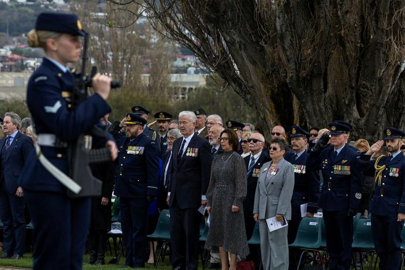 A large group of military personnel and civilians stand up from their chairs.