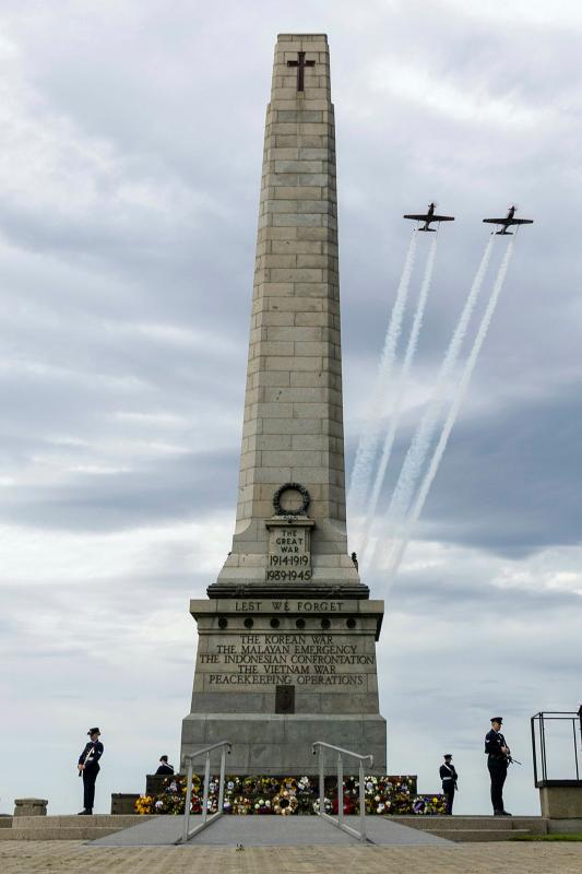 Two aircraft fly past a pillar memorial stationed with four military personnel.