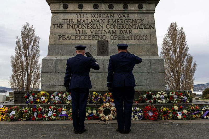Two military men salute a collection of wreaths at the base of a memorial for several wars/operations.