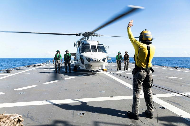 A man in yellow holds up his hand to a helicopter and its crew on the back of a ship.