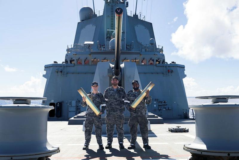 Three Navy officers stand on the bow of a Navy ship holding gun shells.