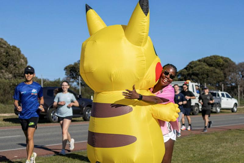 A yellow inflatable cartoon mascot is embraced by a person with other people seen walking on a running track.