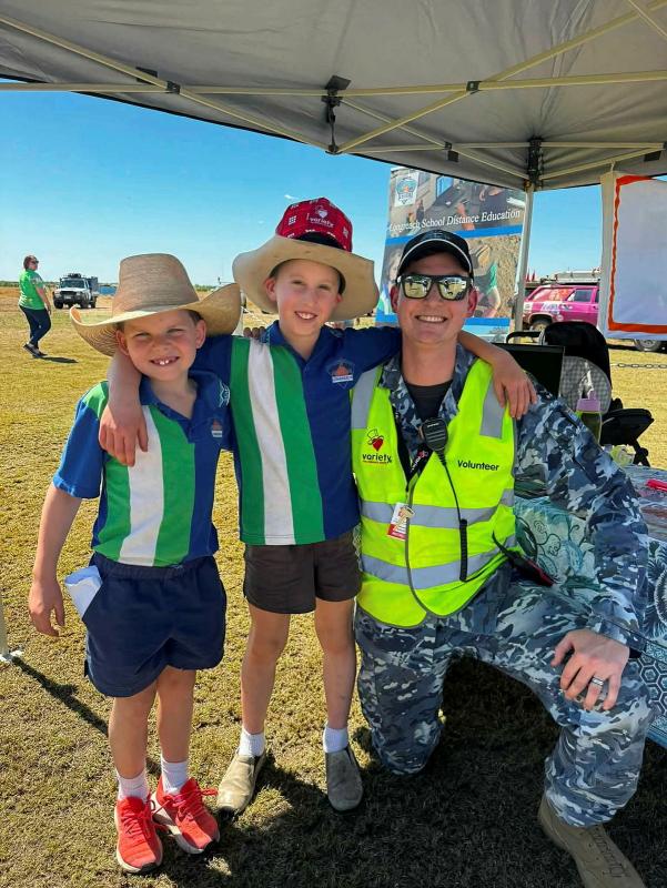 A man in uniform and a high-visibility vest kneels on grass beside two young boys wearing broad-brimmed hats.