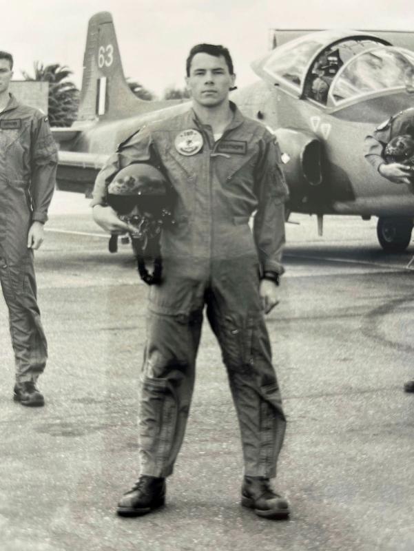 A black-and-white photo of a military pilot holding his helmet before a stationary aircraft.