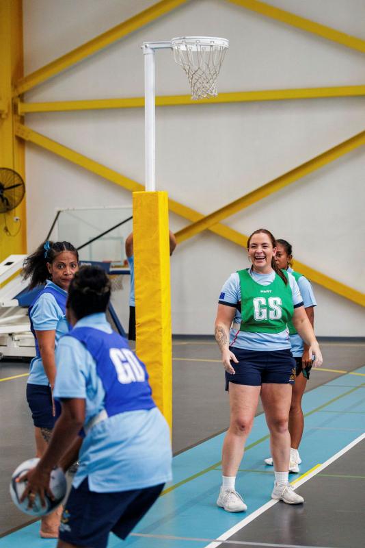 A small group of women face each other near a netball hoop. One has a ball and the others watch her.
