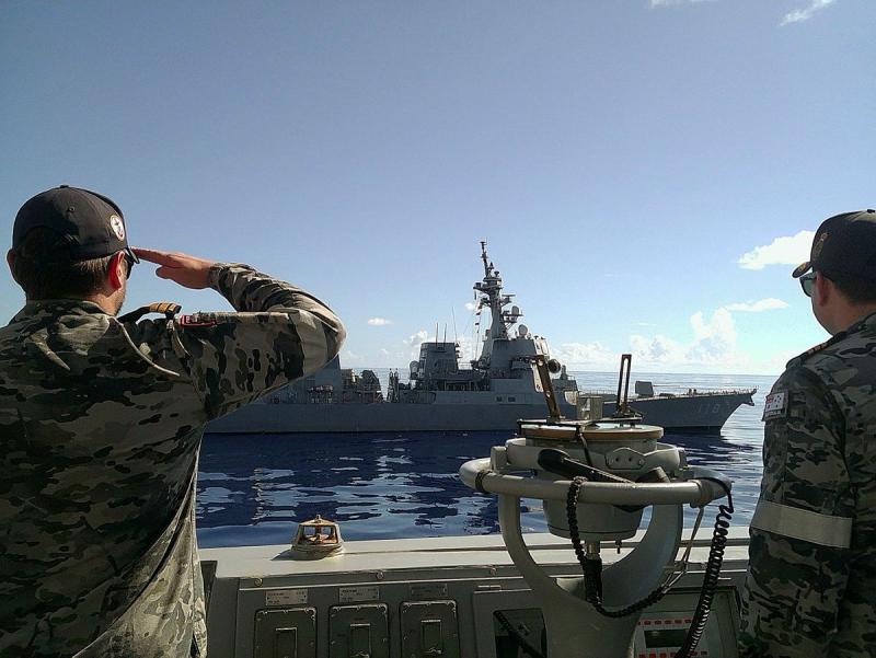 A man in uniform salutes as he faces a ship in the water. Another man in uniform looks on.