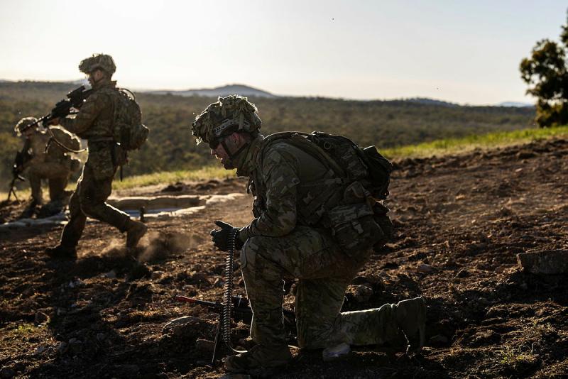 A man in uniform crouches in the dirt on a hill with an ammunition belt draped over his arm and a rifle by his side while other people in uniform run down the hill carrying rifles.