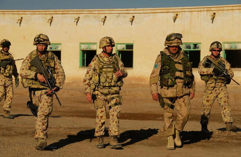 Four people in uniform walk across dirt ground in front of a sand-coloured building, two holding guns.