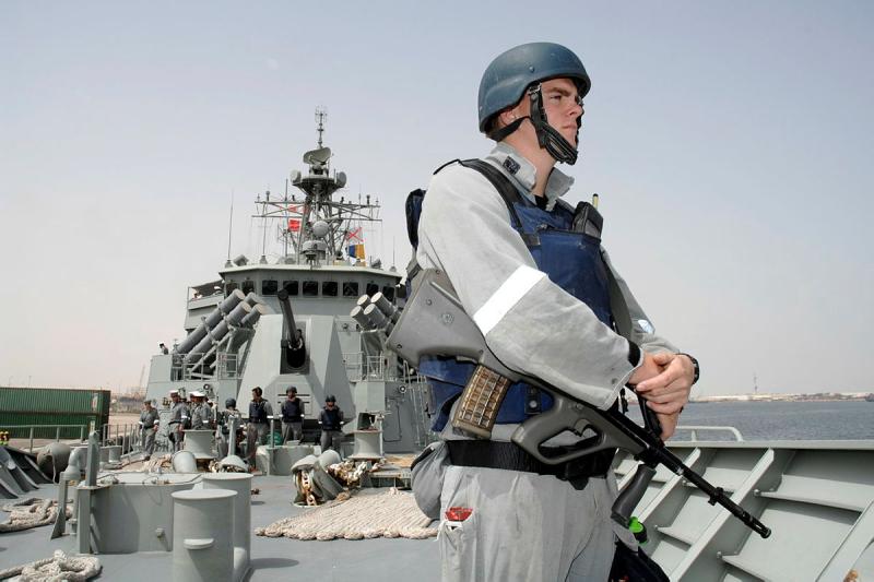 A man in uniform stands on the deck of a ship with a gun hanging from his shoulder.