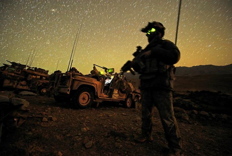 A soldier holding a weapon stands near military vehicles beneath a star-filled sky.