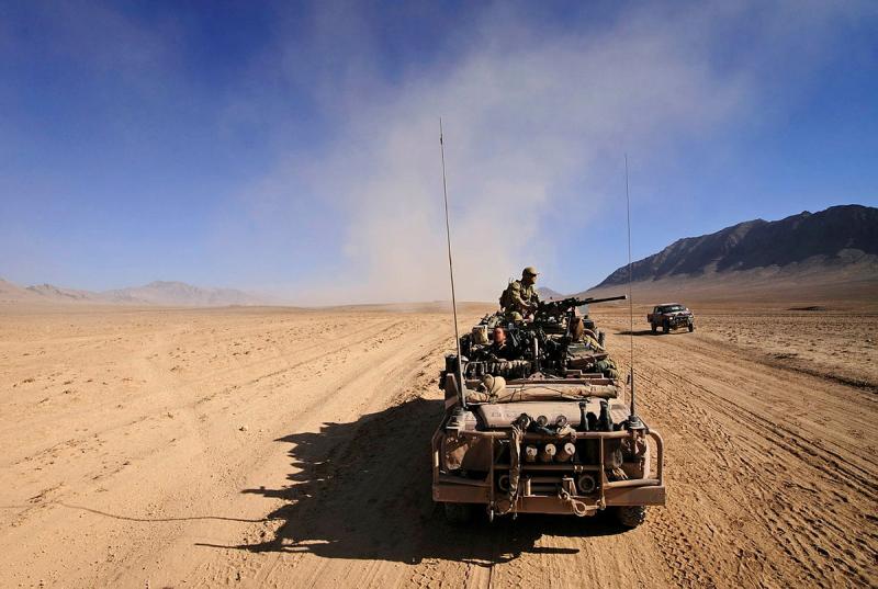 A military vehicle travels through the desert with armed soldiers riding on top. 