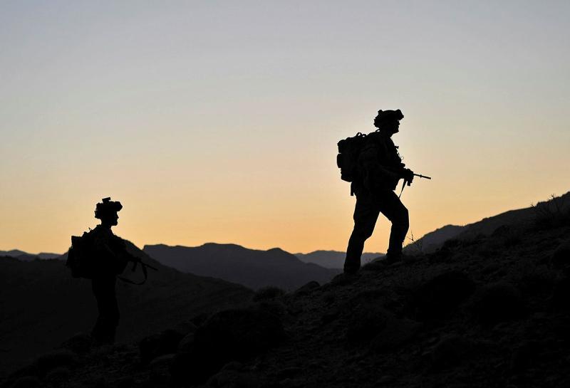 A silhouetted soldier carrying a weapon leads a second figure up a steep hillside against an orange sky.