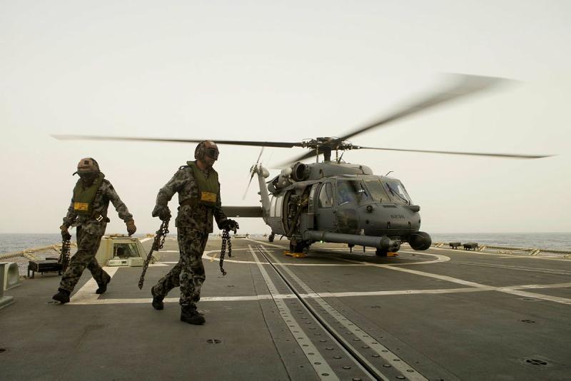 Two military personnel stand on the flight deck of a ship next to a helicopter.