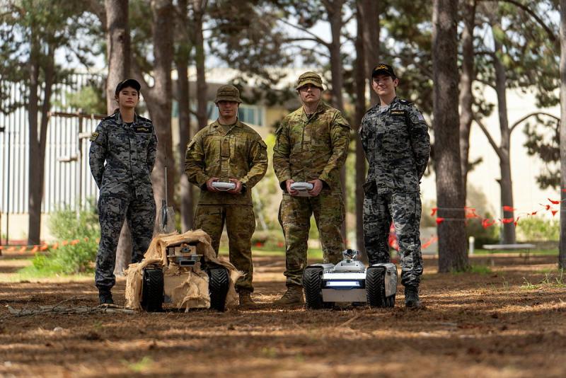 Four military personnel stand with uncrewed ground vehicles. 