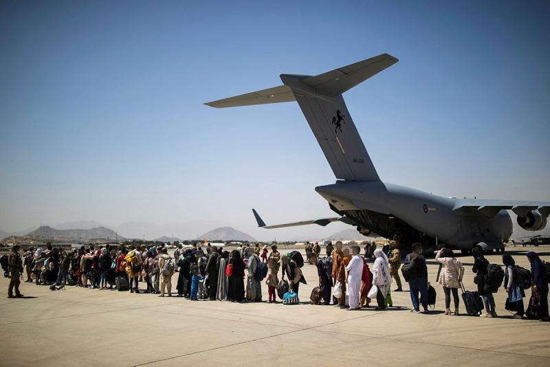 A long line of people stand on an airport tarmac near a plane that has its ramp down