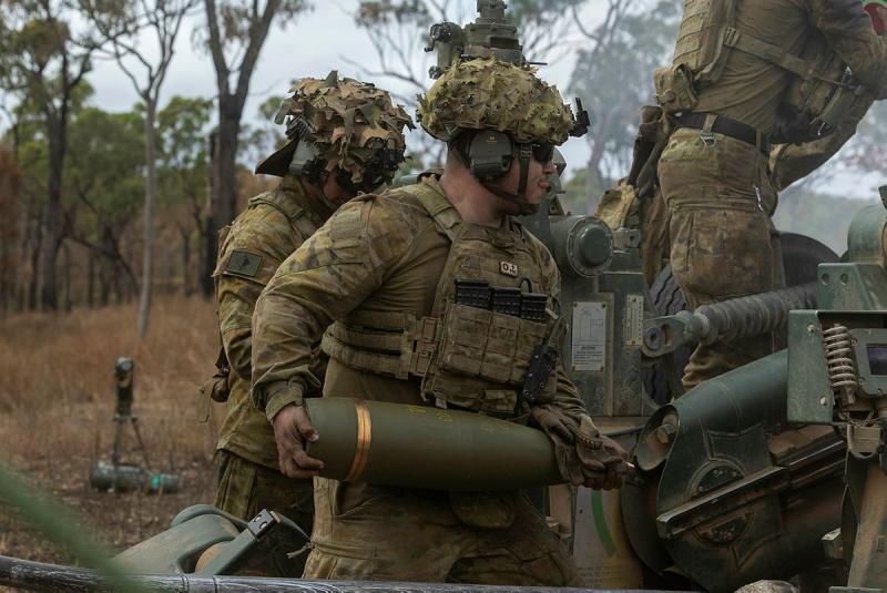 A soldier, surrounded by others in uniform, holds a large artillery shell.
